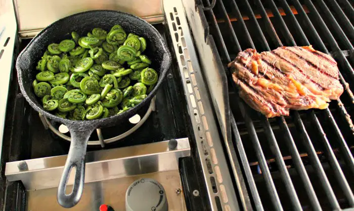 Sauteed fiddleheads cooking in cast iron skillet on gas grill side burner next to seared rib eye steak