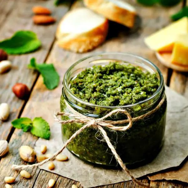 Jar of green pesto sitting on a wooden table with pine nuts, pistachios, basil leaves, and Parmesan cheese nearby.