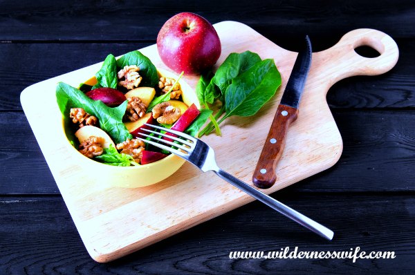 Cutting board with apple, spinach leaves, knife, fork and cream colored bowl full of apple spinach salad.