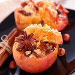 Two stuffed apples on a black plate with some of the spices used in flavoring them. such as cinnamon, star anise, and nutmeg.