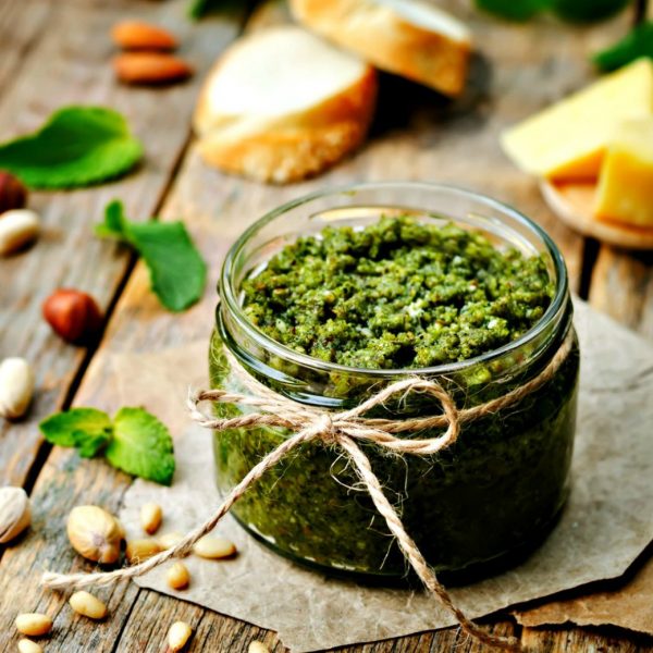 Jar of green pesto sitting on a wooden table with pine nuts, pistachios, basil leaves, and Parmesan cheese nearby.