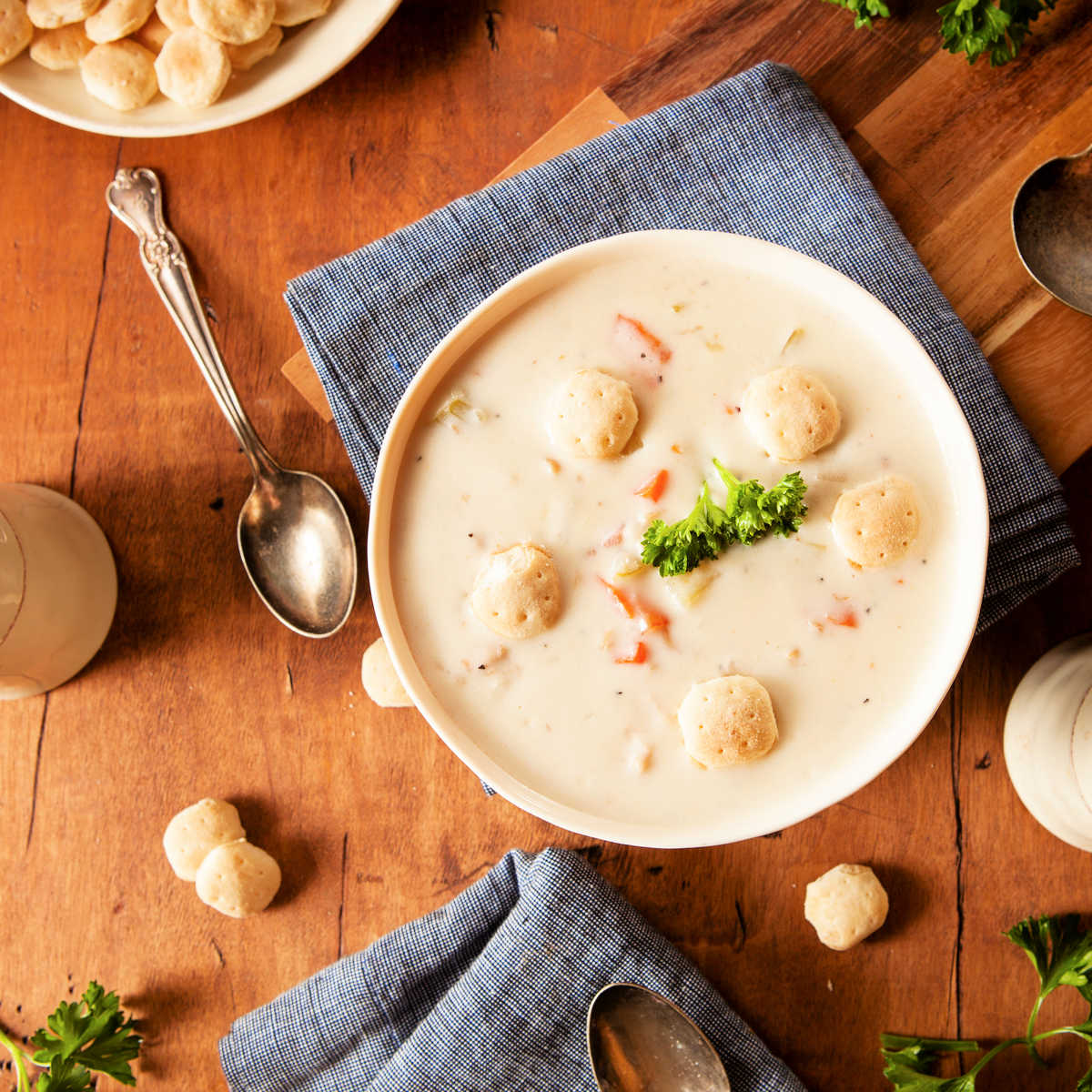 A white bowl set on a blue napkin on a brown wood tabletop. The bowl is filled with clam chowder  crackers floating on and has four soda crackers floating on the surface with a sprig of herbs. 