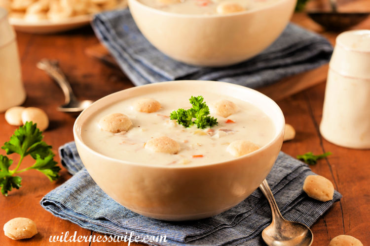 Steaming bowl of New England Clam Chowder set on blue napkin with oyster crackers on wood table