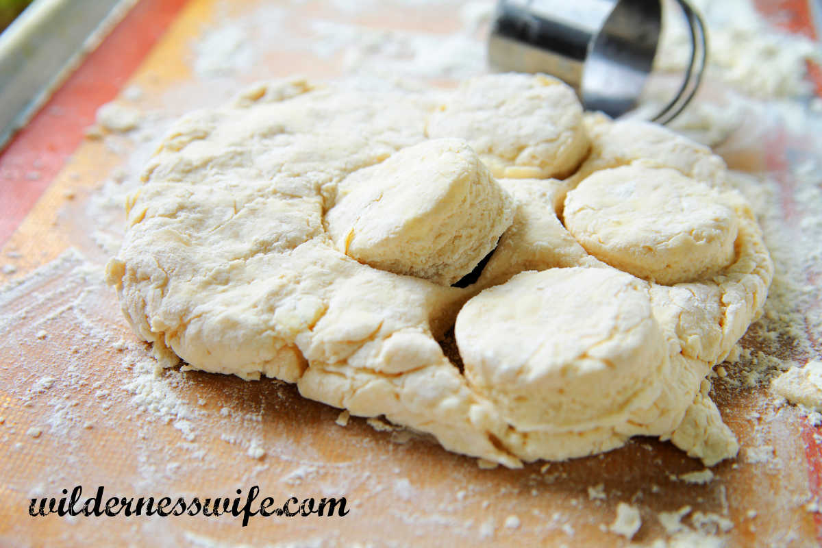 Homemade biscuit dough being cut into individual biscuits on a wooden cutting board