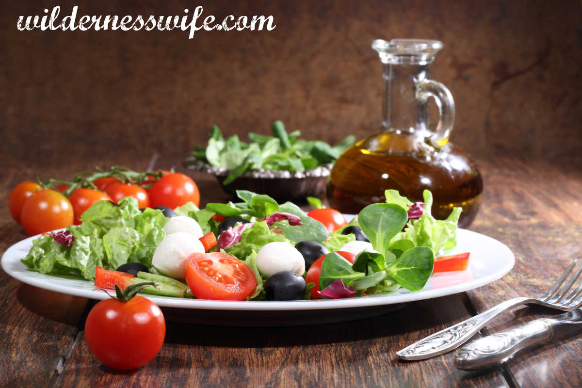 Glass bottle of basic vinaigrette salad dressing set next to a white plate containing salad ingredients including greens and tomatoes.