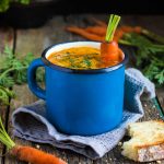 Slow cooker curried carrot soup steaming in a blue mug with a whole carrot sitting upright in the mug as a stirrer. Soup is garnished with chopped green chives. Mug is sitting on a light blue woven napkin next to a slice of crusty fresh baked bread on a wooden table.