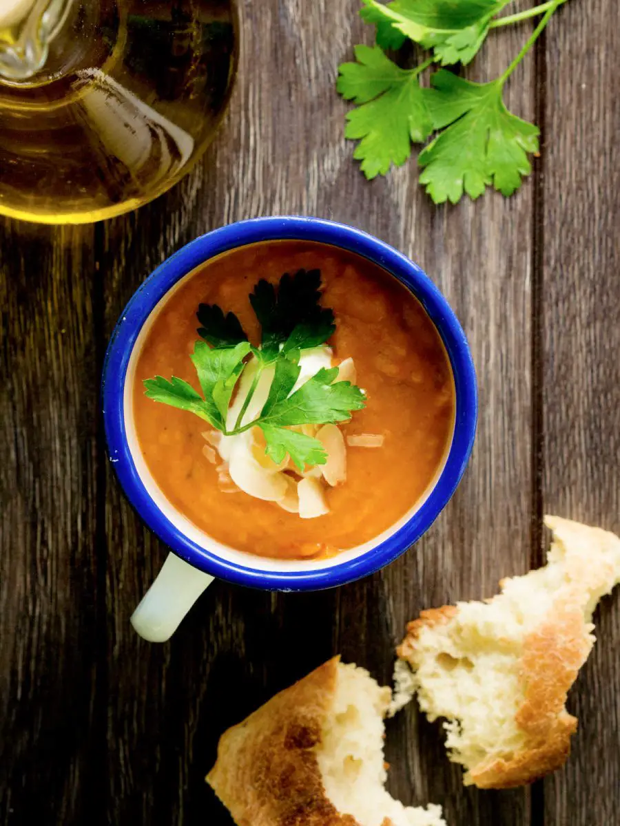 Mug of steaming slow cooker curried carrot soup garnished with a dollop of sour cream and a sprig of flat leaf parsley sitting on a wooden table next to a piece of crusty fresh baked bread. The soup has a spicy curry flavor and yet a sweetness of the carrots.