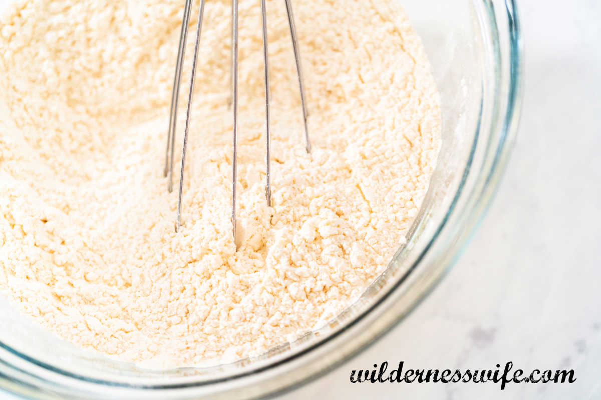 A glass bowl of pancake dry ingredients being mixed with a wire whisk on a light marble counter top