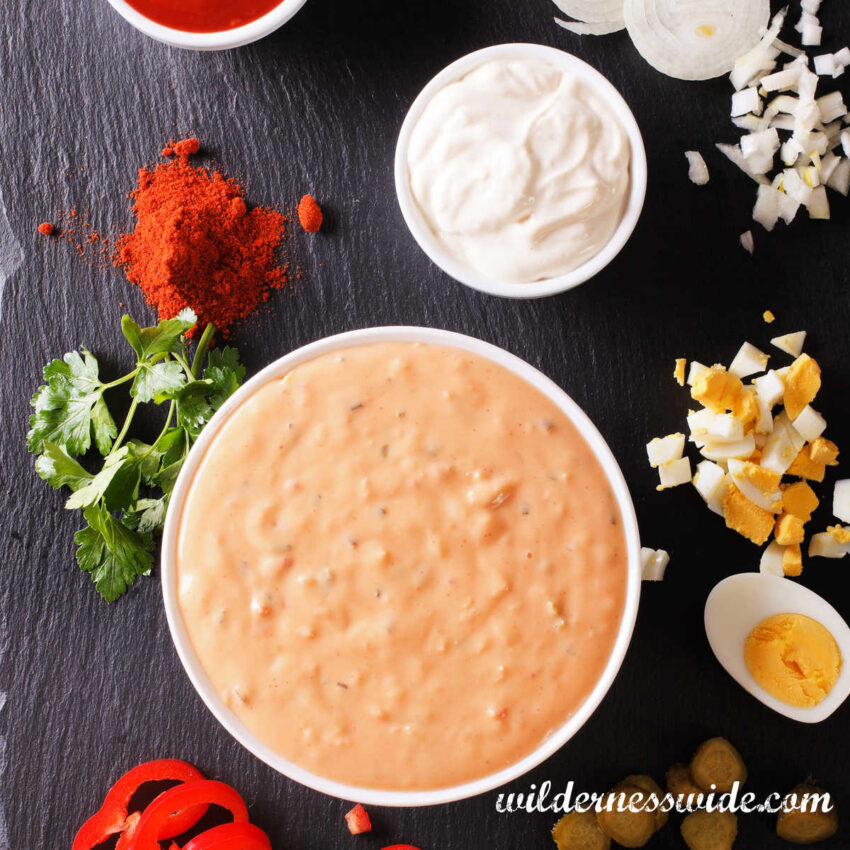 White bowl filled with Russian Dressing set on dark wood table surrounded by necessary ingredients