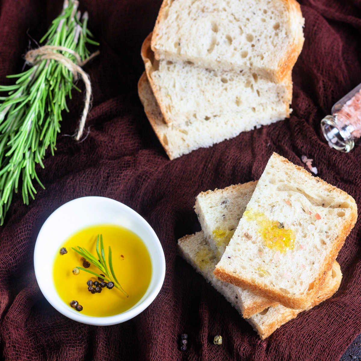 Slices of olive oil and rosemary bread along side a small bowl of olive oil and a  bundle of rosemary