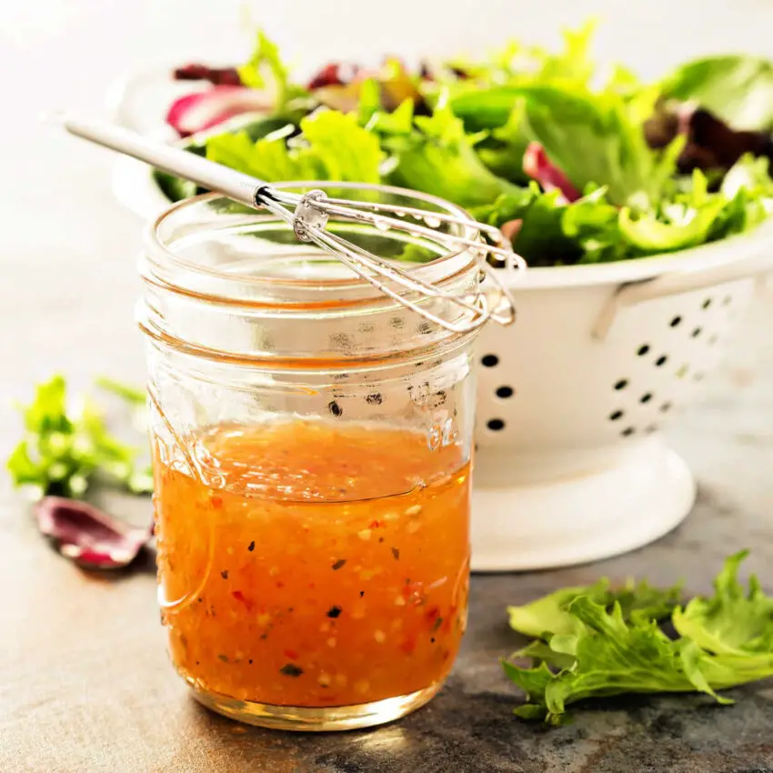 White colander filled with fresh salad greens and canning jar filled with fresh italian dressing with a wire whisk on top
