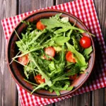 Brown pottery bowl filled with salad comprised of arugula, halved cherry tomatoes, and cucumber slices. The bowl is sitting atop a red and white checked napkin set on a wood table.