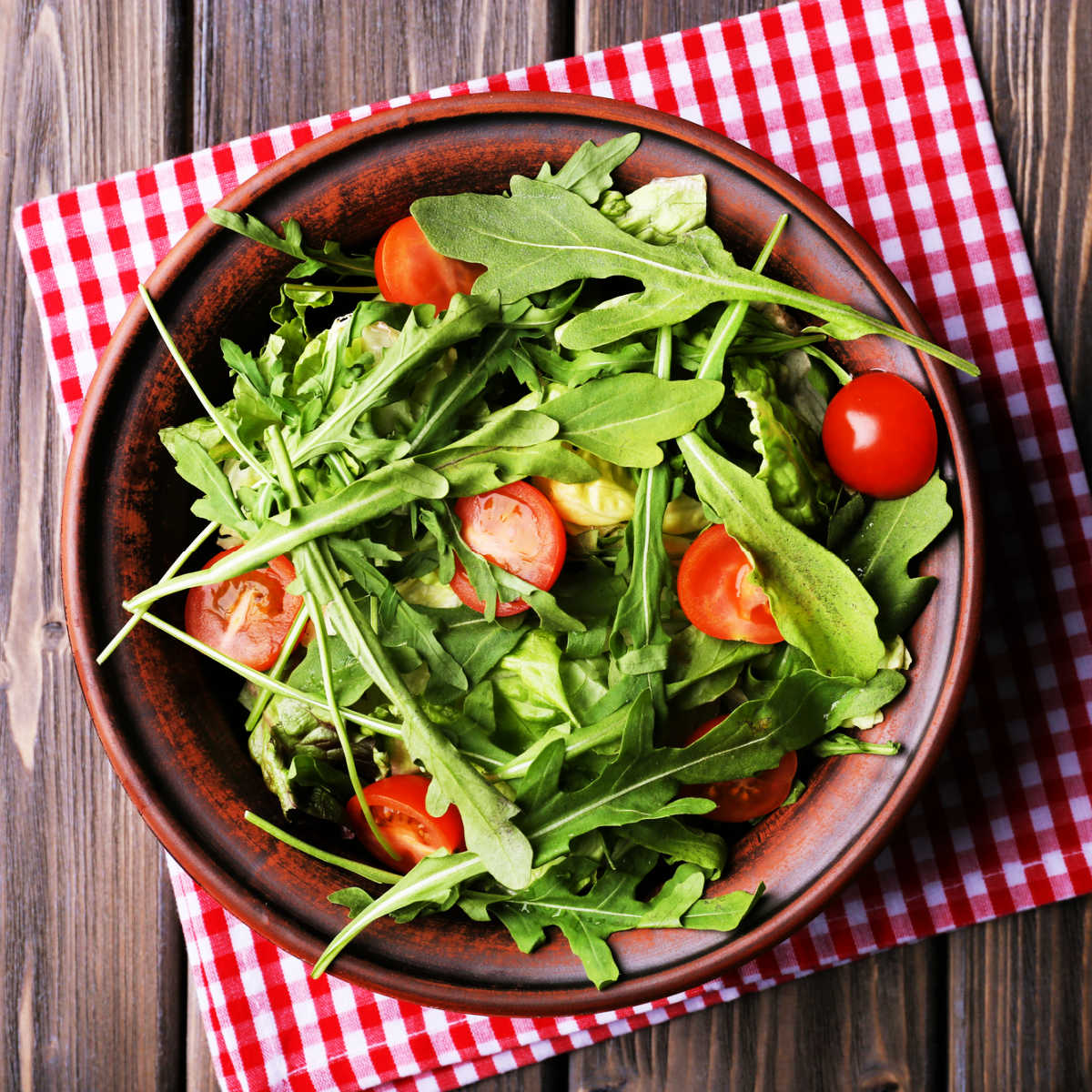 Fresh arugula salad with cherry tomatoes in brown pottery bowl on a red and white checked napkin set on a wood table.