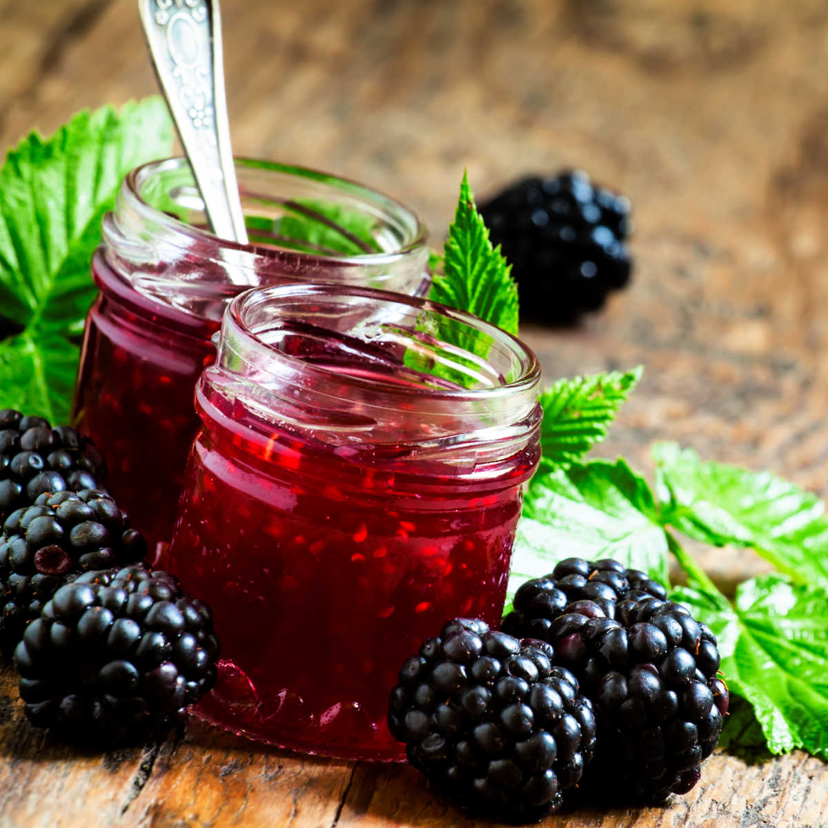 2 jars of blackberry jam surrounded by blackberries on a wood table