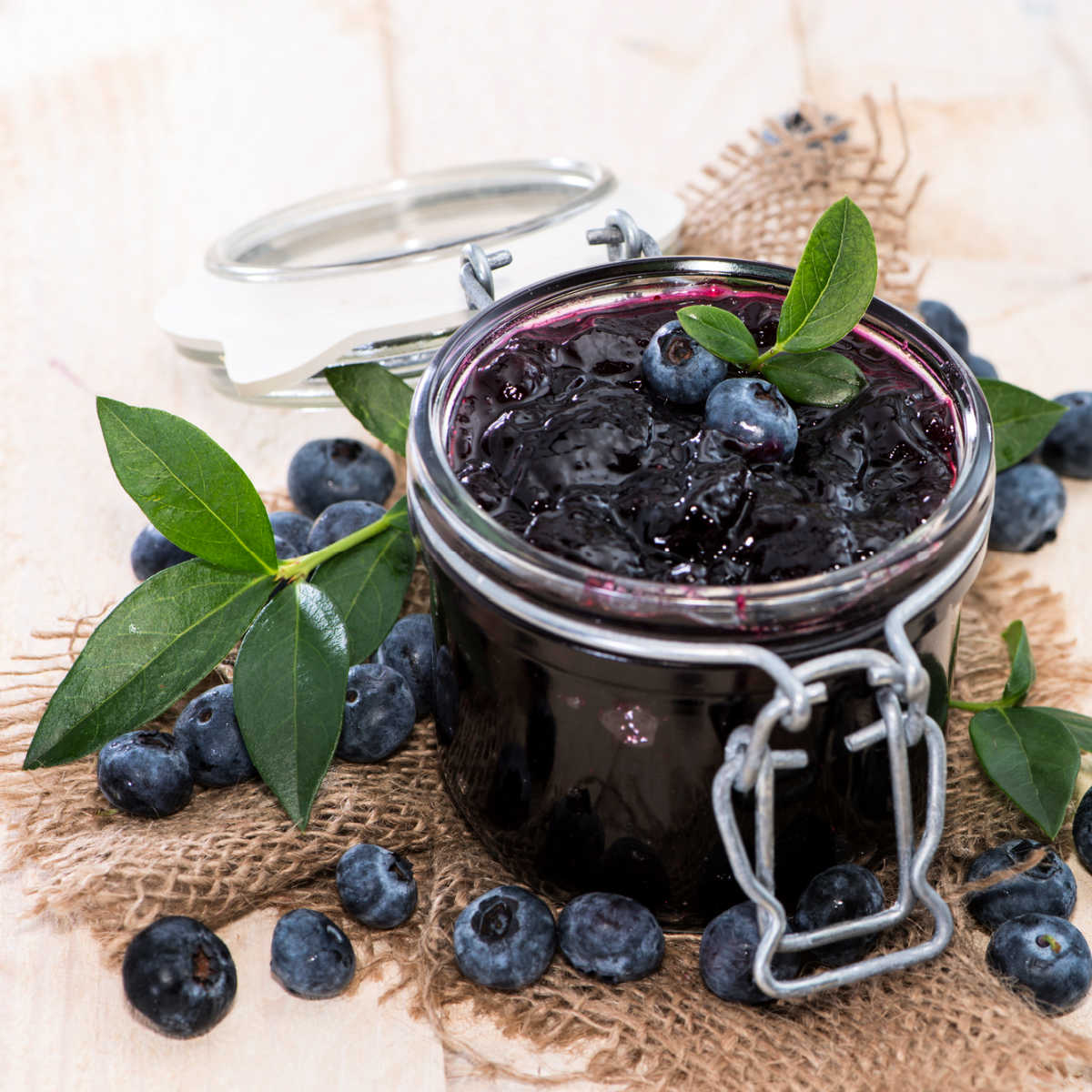 Glass canning jar full of blueberry jam set on a burlap napkin with fresh blueberries setting next to the jar