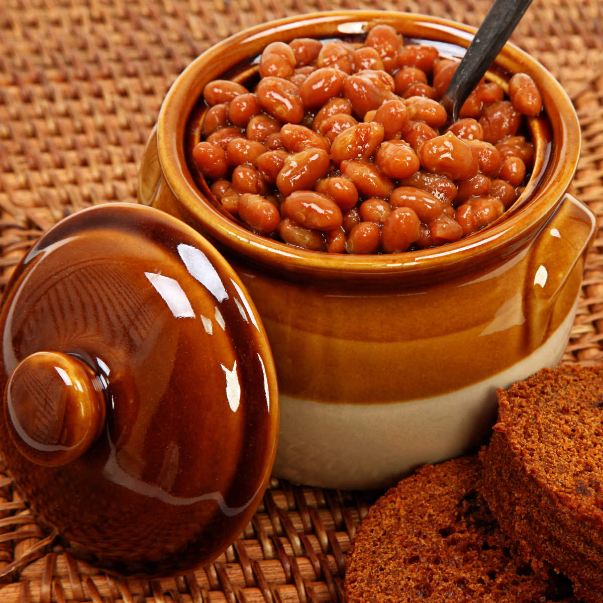 Beanpot full of Boston baked beans with cover leaning against it set on brown woven cloth