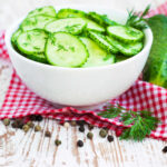 Cucumber salad in a white bowl setting red and white checked napkin on a whitewashed wood table