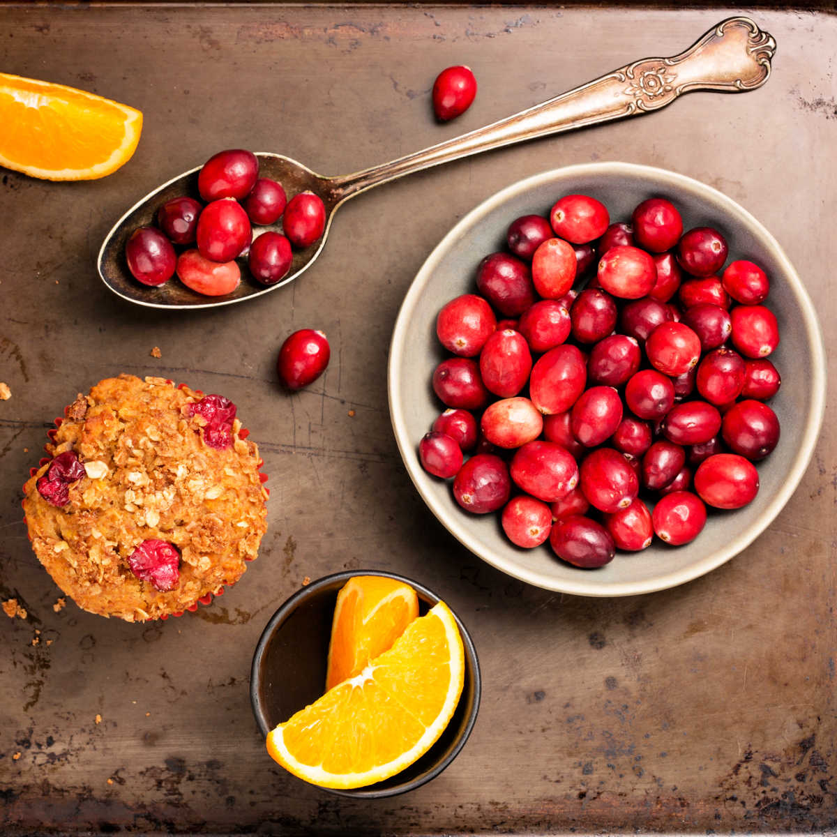 Orange cranberry muffin on a custom dark cement countertop with ingredients called for in recipe - fresh oranges and fresh cranberries.