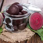 Small glass canning jar containing pickled beets and a raw beet sliced in half sitting on a burlap napkin resting on a wooden table
