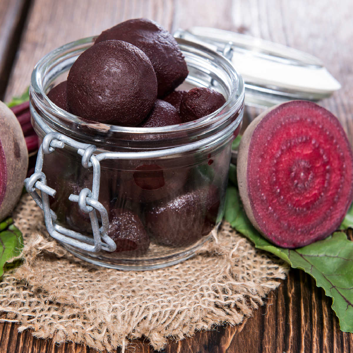 Small glass canning jar containing pickled beets and a raw beet sliced in half sitting on a burlap napkin resting on a wooden table