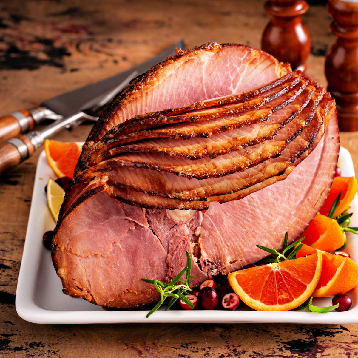 Holiday dinner spiral ham on white platter garnished with fresh cranberry, orange slices, and rosemary sitting on a vintage wooden table.