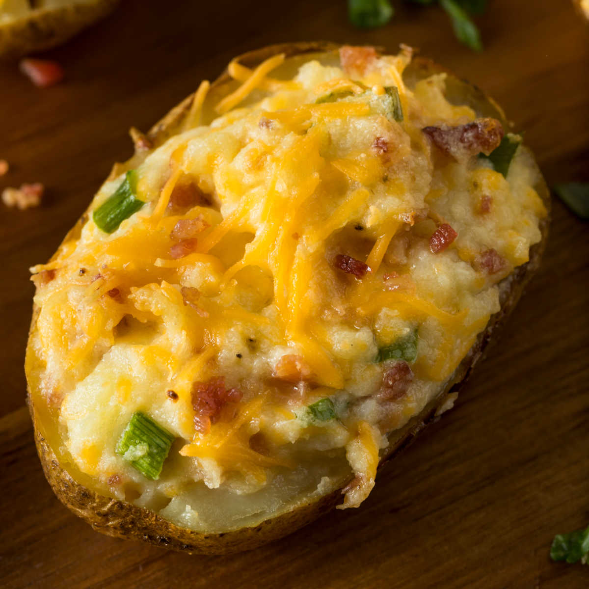 Close-up photo of twice baked potatoes with melted cheese, bacon crumbles, and pieces of chopped chives on wooden cutting board