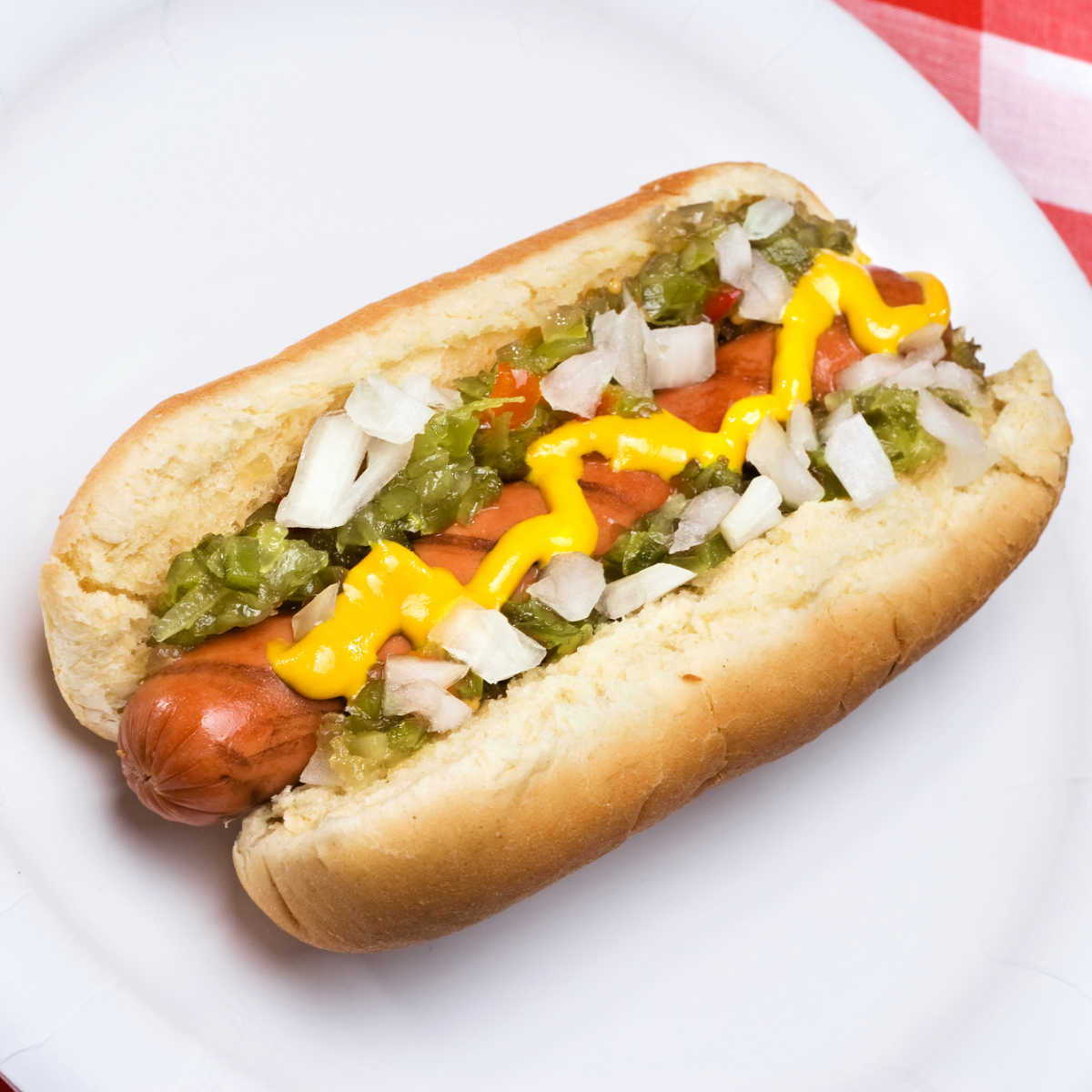 Hot dog in a bun with mustard and zucchini relish and chopped onion on white plate and red checked table cloth