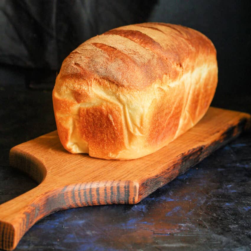 A whole loaf of Anadama bread cooling on a wooden cutting board placed on a black marble countertop