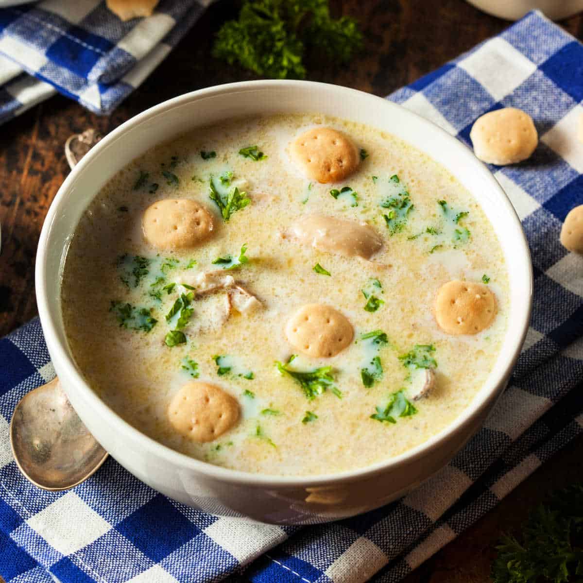 White soup bowl full of New England Clam Chowder with Soda Crackers and fresh chopped parsley as a garnish. The bowl of chowder sits on a blue and white checked napkin on a dark wooden table.