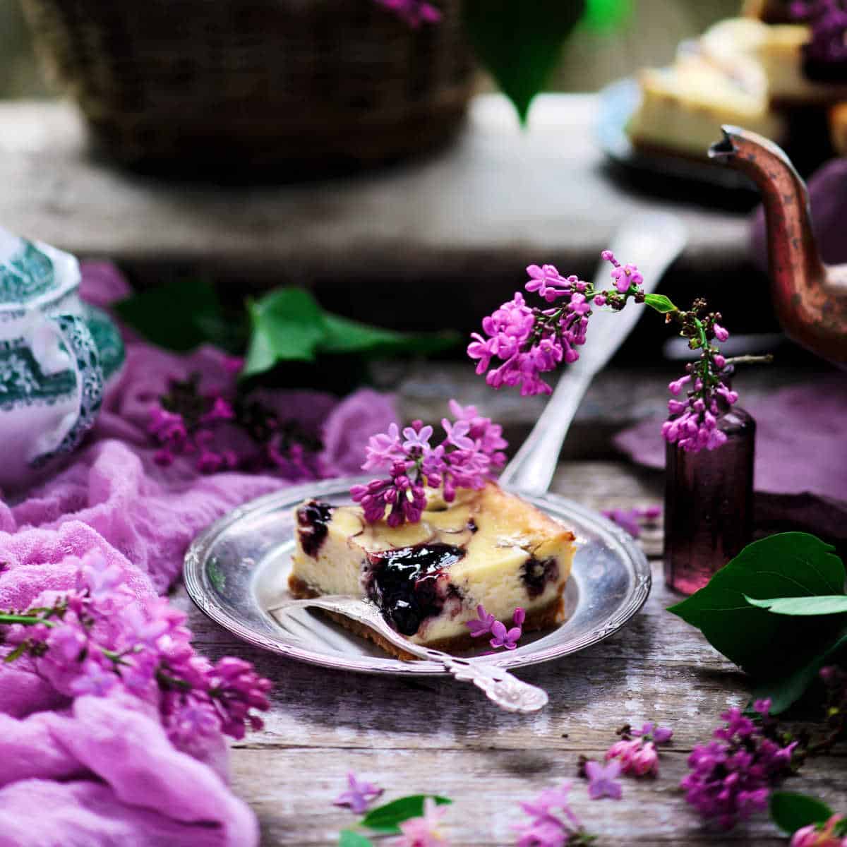 No-bake Blueberry Cheesecake Bar on a grey plate set on weathered wood table with a lavender towel next to it and lilac blossoms scattered around and on it.