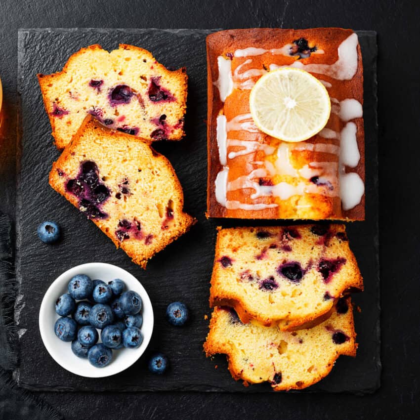 Aloaf of blueberry pound cake on a black slate cutting board with cut slices next to it and a white bowl containing fresh blueberries.