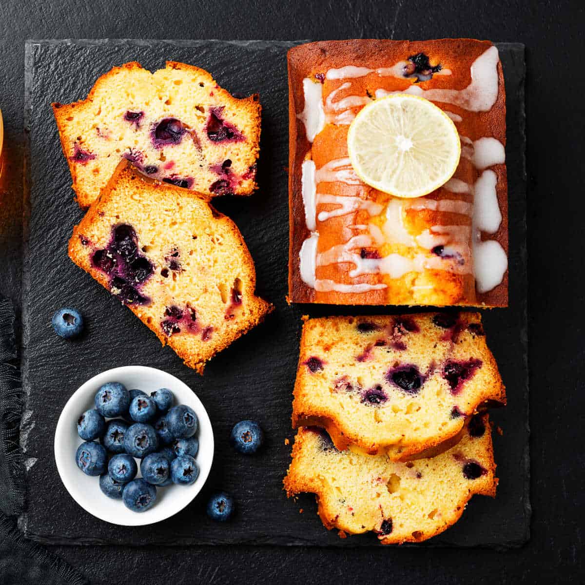 A loaf of blueberry pound cake on a black slate cutting board with cut slices next to it and a white bowl containing fresh blueberries.