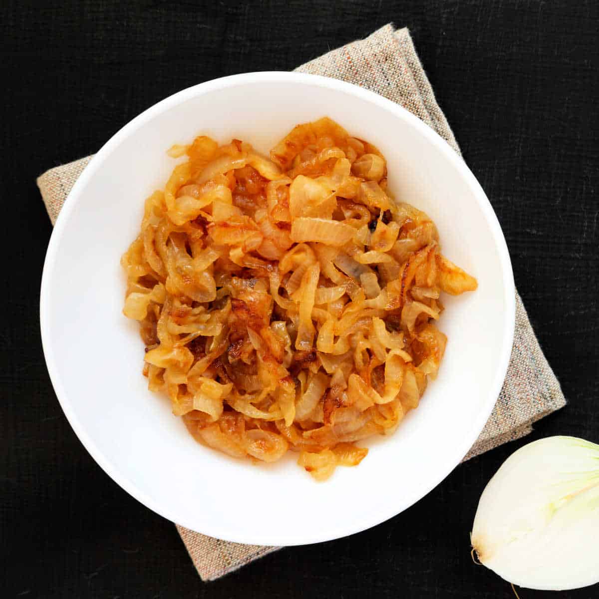 A white bowl filled with golden caramelized onions sitting on a tan burlap napkin on a black granite countertop.