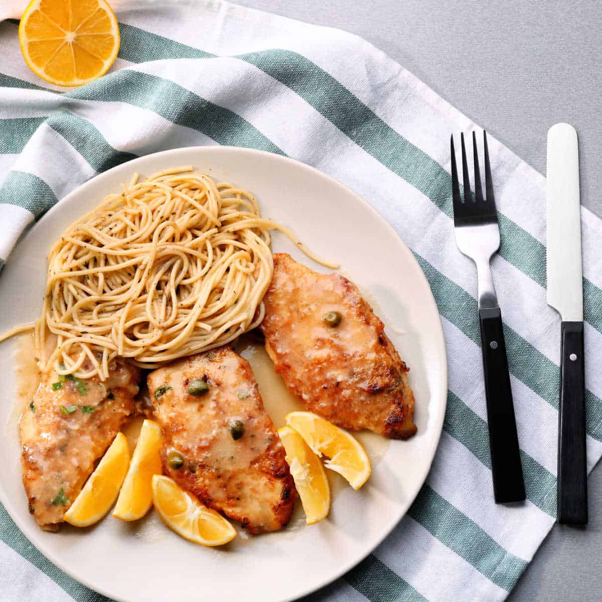 White plate holding Chicken Piccata, pasta and lemon wedges set on a white and green striped napkin. A knife and fork are to the left of the plate.