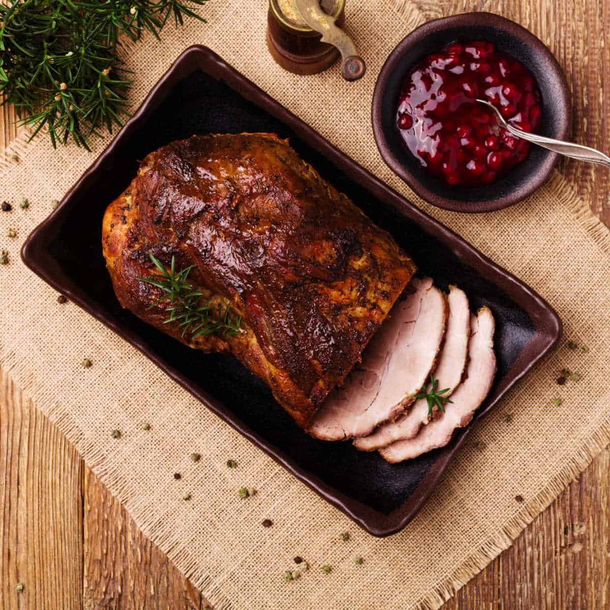 Pork roast cooked in slow cooker glazed with cranberry sauce in roasting pan set on tan burlap napkin with cranberry sauce glaze in a brown bowl