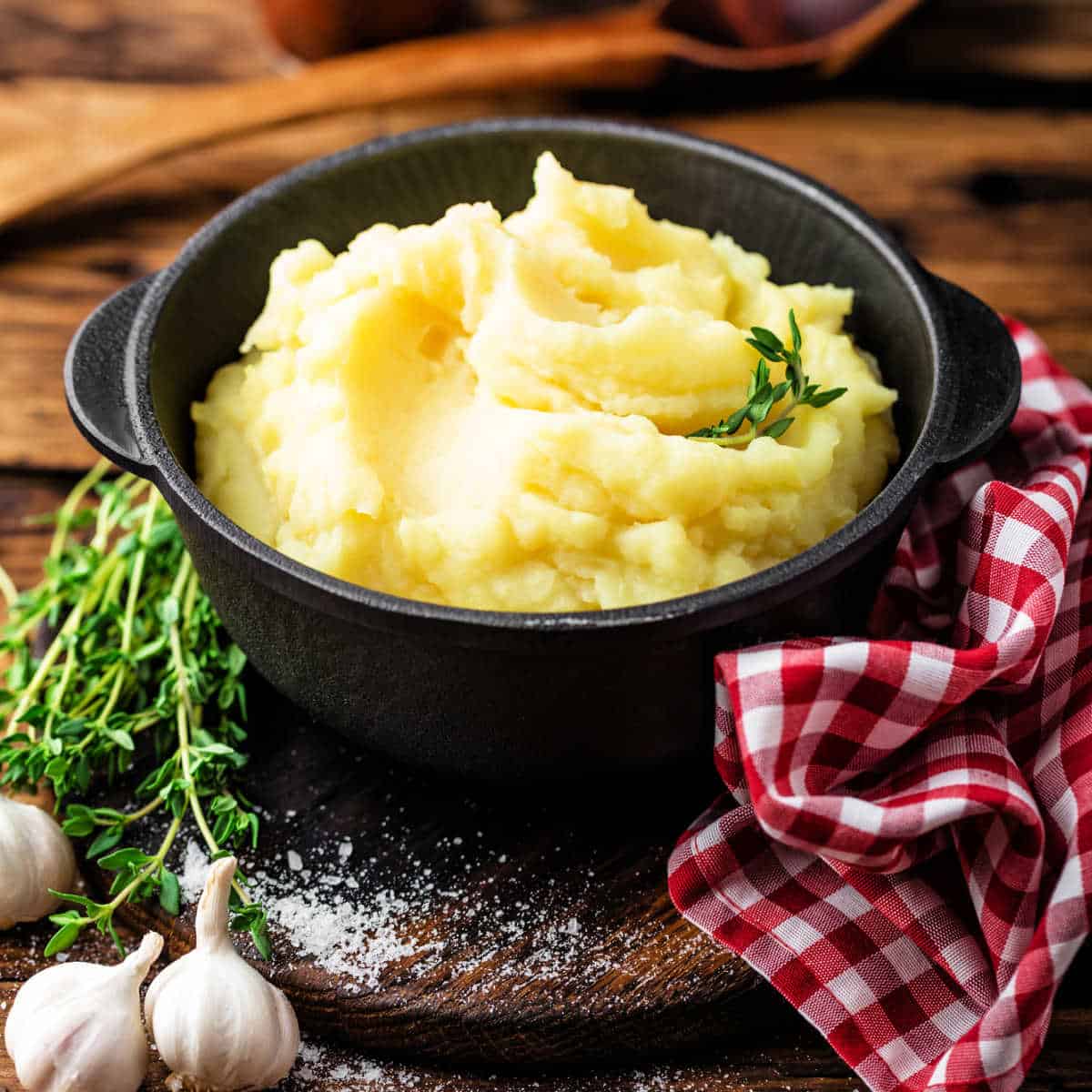 Black serving dish, filled with mashed potatoes, wrapped in a red and white checkered napkin set on a brown wooden table with fresh thyme sprigs and garlic bulbs on the left side.