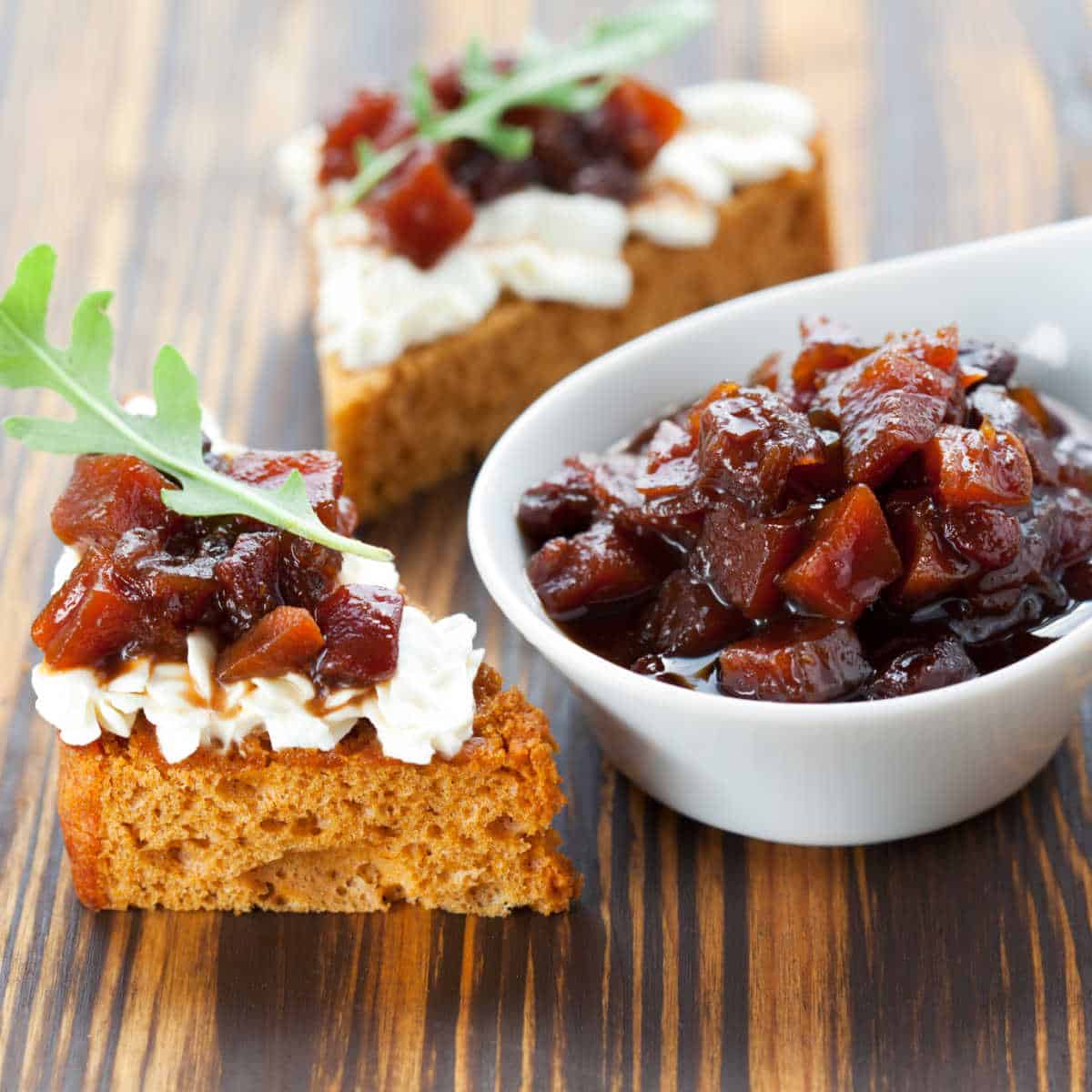 Chunky Pumpkin Chutney in white serving bowl set on brown wooden table. To the left is a piece of pumpkin bread with cream cheese and chutney spread on it.